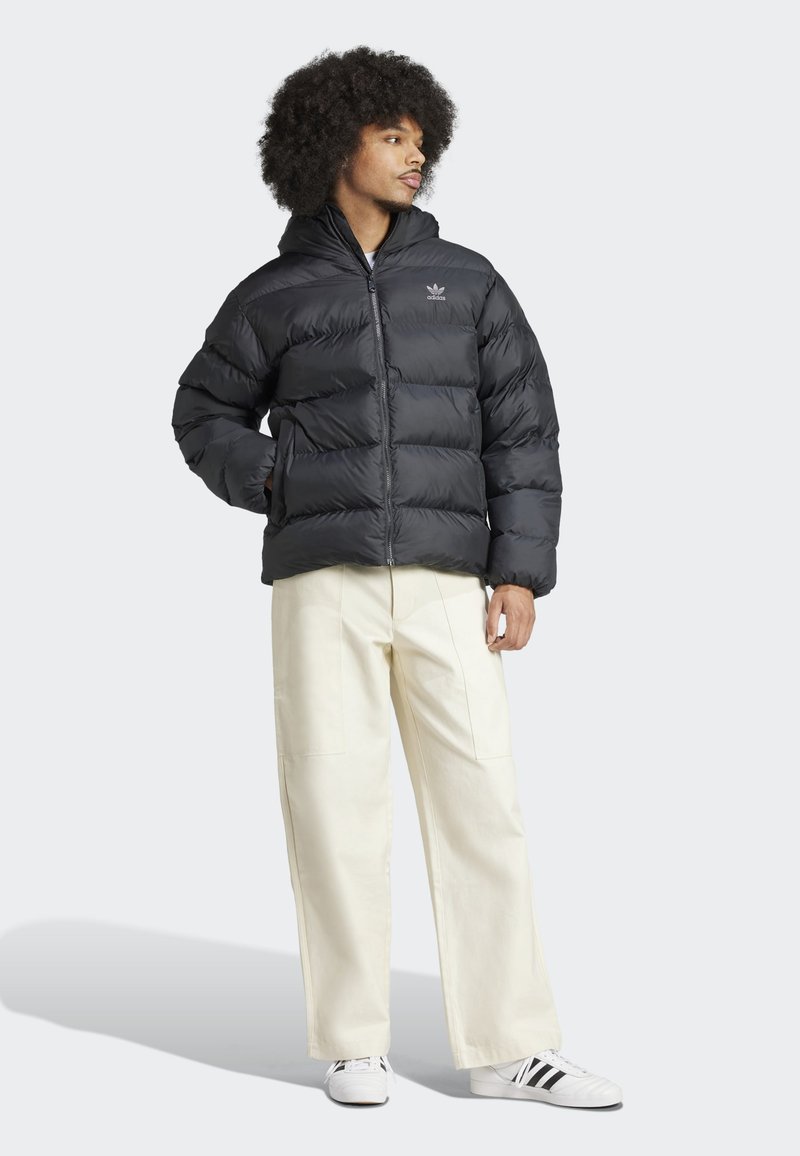 Schwarze Steppjacke mit Reißverschluss vorne, Kapuze und horizontalem Steppmuster. Kombiniert mit cremefarbenen, weiten Hosen und weißen Sneakers.