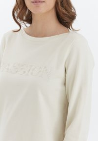 Top de manga larga en color crema con cuello redondo y texto en relieve "PASSION", textura suave y corte relajado.