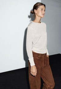 Jasnobeżowa bluzka z długim rękawem w połączeniu z brązowymi zamszowymi spodniami o szerokich nogawkach. Strój charakteryzuje się luźnym krojem i teksturowanym materiałem.