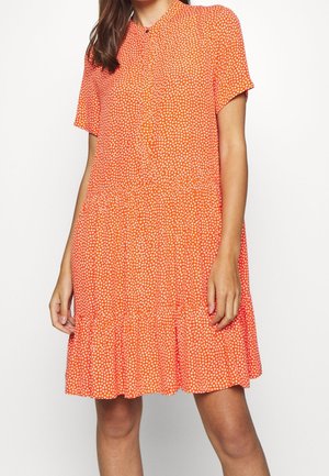 Woman wearing a short-sleeve, button-front orange dress with small white polka dots and a ruffled hem standing against a white background.
