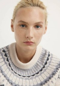 Giovane donna con pelle chiara, occhi blu e capelli biondi raccolti, che indossa un maglione a maglia bianco e grigio con un motivo, mentre guarda dritto davanti a sé.