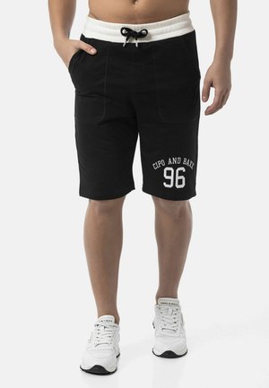 Man draagt zwarte shorts met witte trekkoorden en logo "Cipo and Baxx 96" op het linkerbeen, gecombineerd met witte sportschoenen.
