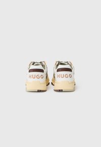 Sport shoes with a white and olive upper, brown accents, and a beige sole; features "HUGO" logo prominently on the back.