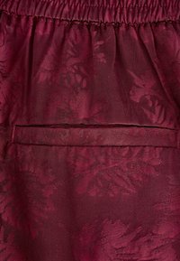 Bordeaux stof met een gestructureerd bloemenpatroon, voorzien van een elastische tailleband en een discreet ingezette zak op de rug.