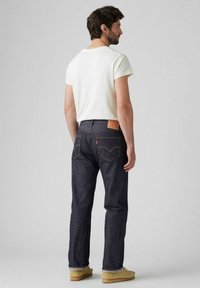 Jeans de mezclilla azul oscuro con un corte recto, acentuados con costuras amarillas y una etiqueta de cuero marrón en la cintura. Camiseta blanca, zapatos beige.