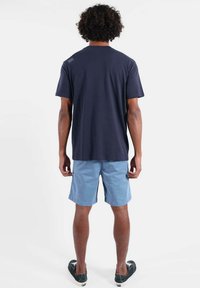 T-shirt en coton bleu marine avec col rond, manches courtes, et un petit détail de design dans le dos, assorti à un short bleu clair et des chaussures noires.