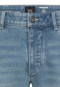 Close-up of light blue denim jeans waist with button, zipper, belt loops, and branded inner label reading "Camel Active" and "Fit Houston."