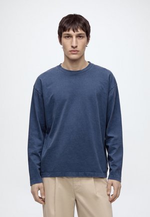Un homme porte une chemise bleu marine à manches longues et un pantalon beige sur un fond clair et uni.