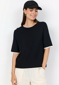 Kvinna som ler med stängda ögon, iförd svart keps, svart kortärmad topp med vit kant och off-white byxor, som står mot en vit bakgrund.