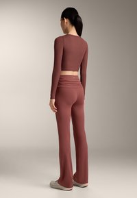 Donna con capelli scuri raccolti in una coda di cavallo, indossa un top corto aderente a maniche lunghe color ruggine e pantaloni a vita alta svasati abbinati, con scarpe da ginnastica bianche.