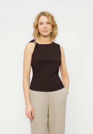 Femme blonde portant un haut sans manches noir texturé avec détail anneau métallique et un pantalon beige, debout avec une main dans la poche.
