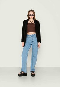 Blazer preto sobre um top cropped castanho, combinado com jeans azul claro e sandálias de plataforma pretas e grossas. Acessórios incluem óculos de sol escuros.