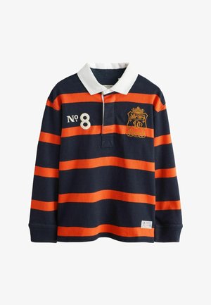 Blond dieťa v námornícko-modro-oranžovo pruhovanom rugby tričku cez denimovú košeľu, béžové nohavice, ruky v vreckách, stojaci oproti jednoliatej bielej stene.