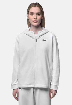 Femme portant un sweat à capuche blanc zippé et un pantalon blanc assorti avec des logos noirs Kappa, debout face à l'avant sur un fond uni.