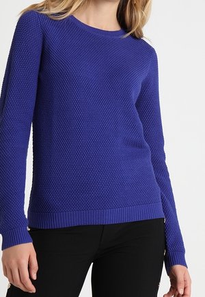 Pull en tricot de couleur bleu vif avec un motif texturé en nid d'abeille. Encolure ronde et poignets et ourlet côtelés. Porté avec un pantalon noir.