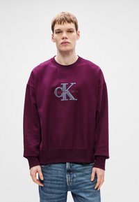 Bordový oversize svetr s kulatým výstřihem, se stříbrným logem a textovým designem. Texturovaná tkanina s žebrovanými manžetami a lemem.