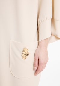 Beige Kleidungsstück mit weiten Ärmeln und einer Tasche, die mit einem goldenen, strukturierten Akzent verziert ist. Glattes, weiches Material mit einem dezenten Glanz.