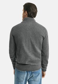 Grauer gestrickter Pullover mit hohem Kragen, gerippten Bündchen und Saum, über hellblauen Jeans getragen, von hinten fotografiert.