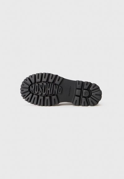 Semelle en caoutchouc noir avec des rainures profondes et des détails texturés. Le logo "MOSCHINO" est embossé au centre, offrant un design durable et structuré.