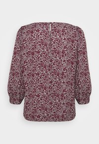 Blouse à manches longues avec un motif floral bordeaux sur un tissu gris clair, manches bouffantes et une petite ouverture en forme de goutte dans le dos avec fermeture par bouton.