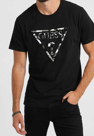 Homme portant un t-shirt noir Guess avec un logo triangulaire monochrome et un pantalon noir, une main dans la poche et l'autre détendue à ses côtés.
