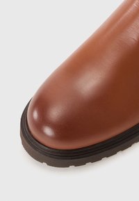 Brown leather boot with a rounded toe and a smooth texture, featuring a thick black rubber sole with layered detailing.