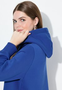 Niebieska bluza z kapturem o miękkiej teksturze, wyposażona w sznurek przy kapturze, ściągacze na mankietach i luźny krój. Tło to jednolita biała ściana.