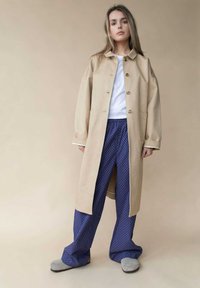 Beige langer Mantel mit Knöpfen und zwei Taschen, getragen über einem weißen Hemd und navyblauen gepunkteten Hosen. Graue Hausschuhe runden den Look ab.