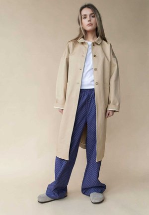 Beige lange jas met knopen en twee zakken, gedragen over een wit shirt en marineblauwe broek met stippen. Grijze slippers maken de look compleet.