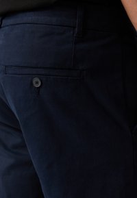 Gros plan sur un pantalon bleu marine foncé avec une poche arrière fermée par un seul bouton noir, porté par une personne.