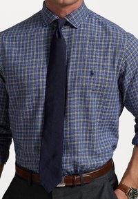 Blå och vit rutig skjorta med spridd krage, ihop med en mörk marinblå slips. Anmärkningsvärd broderad logotyp på bröstet.