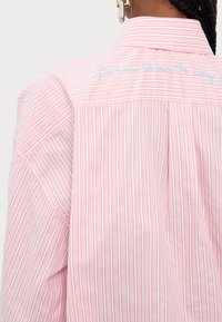 Camisa em riscas cor-de-rosa com uma parte traseira com bordado. O tecido é leve e suave, com riscas finas e contrastantes.