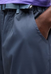 Χέρι με σκούρο δέρμα που ακουμπά στην τσέπη ναυτικού μπλε παντελονιού με ελαστική μέση και μπλε ζώνη, φορώντας μοβ πουκάμισο κρυμμένο μέσα.