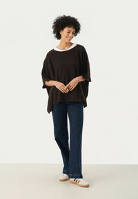 Bruine oversized gebreide top met een ronde halslijn, gelaagd over een wit shirt. Gecombineerd met donkere denim broeken en witte sneakers.