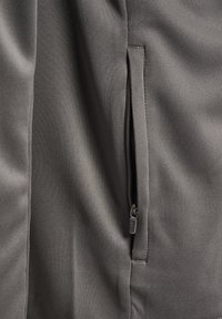 Nahaufnahme eines grauen Stoffkleidungsstücks mit einer leicht geöffneten, vertikalen Reißverschlusstasche, die den Reißverschlussschieber zeigt.