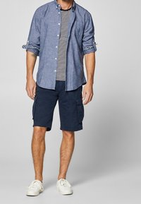 Chemise en chambray bleu, t-shirt rayé blanc et marine en dessous, shorts cargo navy, et baskets blanches. Tenue décontractée avec les manches retroussées.