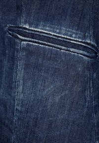 Dunkles Denim-Gewebe mit einer Nahaufnahme einer kleinen, schrägen Tasche, die subtile Stichdetails und eine leicht verblasste Textur aufweist.