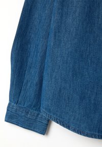 Camicia di denim con un ricco colore blu, texture morbida, polsini con bottoni e orlo curvo. Dettaglio di cuciture visibili lungo i bordi.