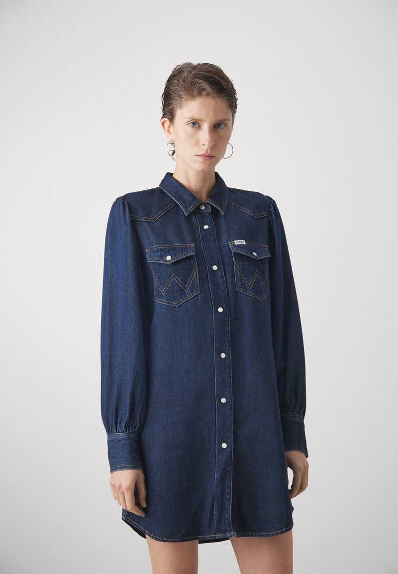 Robe chemise en denim avec de longues manches bouffantes, devant boutonné, deux poches poitrine et couture contrastante en bleu foncé.