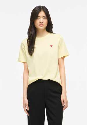 Jeune femme aux longs cheveux foncés portant un t-shirt jaune pâle avec un petit cœur rouge sur la poitrine et un pantalon noir sur fond blanc.