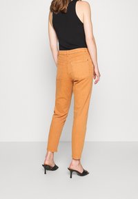 Jeans de talle alto color beige con un corte cónico y bolsillos traseros, combinados con mules negros de tacón y una blusa negra sin mangas.