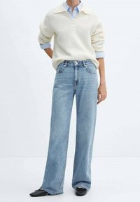 Persona con un suéter de punto crema sobre una camisa de botones azul, jeans de tiro alto y pierna ancha de color azul claro, y bailarinas negras, de pie frente a un fondo blanco.