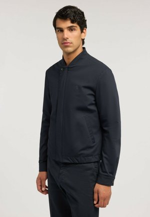 Uomo con capelli scuri e ricci, indossa una giacca blu scuro e pantaloni abbinati, in piedi davanti a uno sfondo chiaro e semplice.