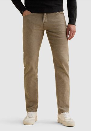 Homme portant un pantalon beige en velours côtelé, des baskets blanches et un t-shirt noir à manches longues, une main dans la poche, sur un fond uni.