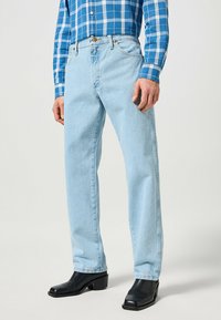 Jeans en denim bleu clair avec un design à jambe droite, cinq poches et une légère décoloration, portés avec une chemise à carreaux bleus et des bottes noires.