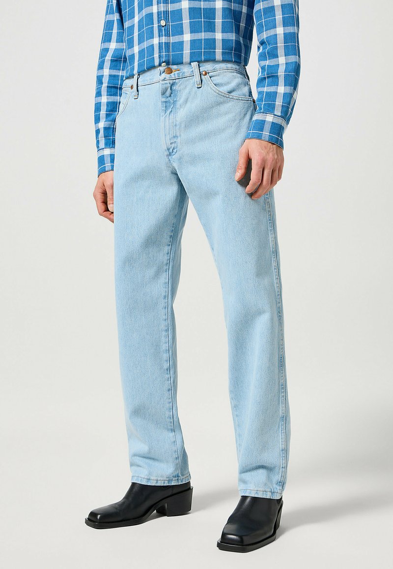 Jeans en denim bleu clair avec un design à jambe droite, cinq poches et une légère décoloration, portés avec une chemise à carreaux bleus et des bottes noires.