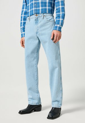 Homme portant un jean bleu clair droit, des bottes noires à bout carré et une chemise à carreaux bleue à manches longues, debout devant un fond clair.