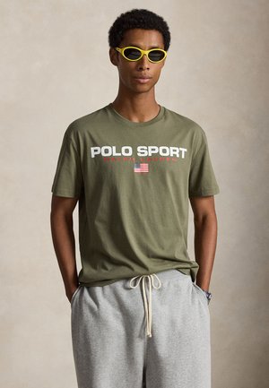 Junger Mann mit gelber Sonnenbrille, olivgrünem Polo Sport Ralph Lauren T-Shirt mit amerikanischer Flagge und hellgrauer Jogginghose mit Kordelzug.