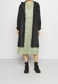 Musta vedenpitävä takki hupulla ja joustavilla hihansuilla vihreän kukkamekon päälle, jossa on röyhelöyksityiskohtia, yhdistettynä mustiin nilkkureihin.