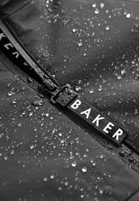 Detail čiernej nepremokavej tkaniny s kvapkami vody na jazdci zipsu označenom nápisom "BAKER" bielymi písmenami.